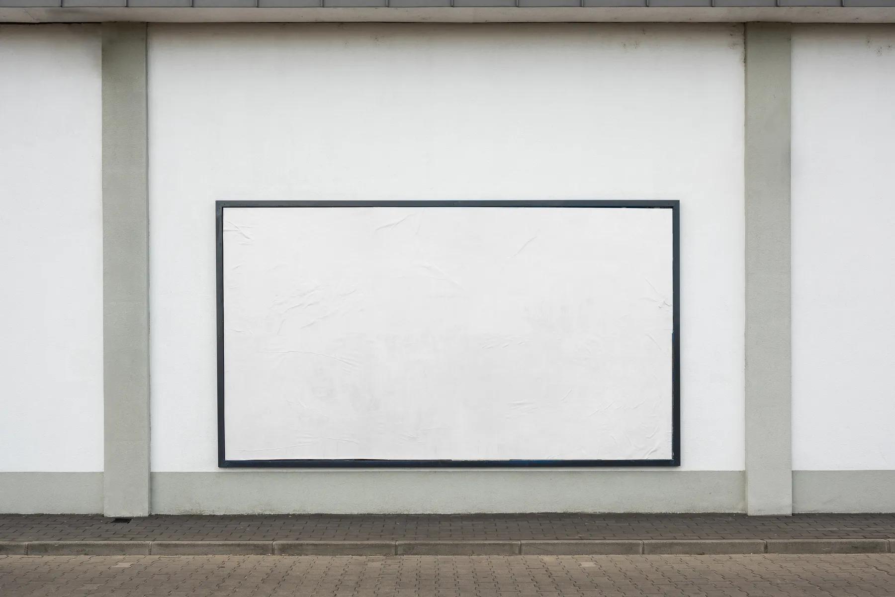 Clean and empty billboard mockup mounted on a white building facade with a grey roof and paved ground in front, suitable for graphic designers to display their advertising work.