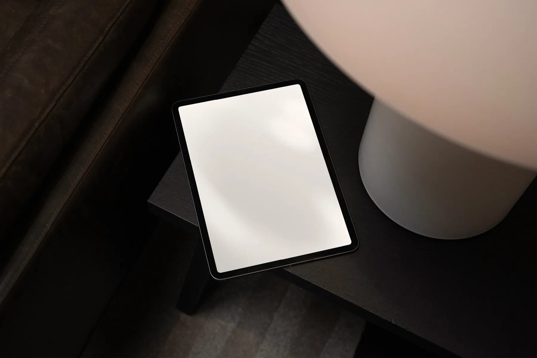 Top view mockup of a blank tablet screen placed on a black table beside a large white lamp, creating a modern, minimalistic scene.