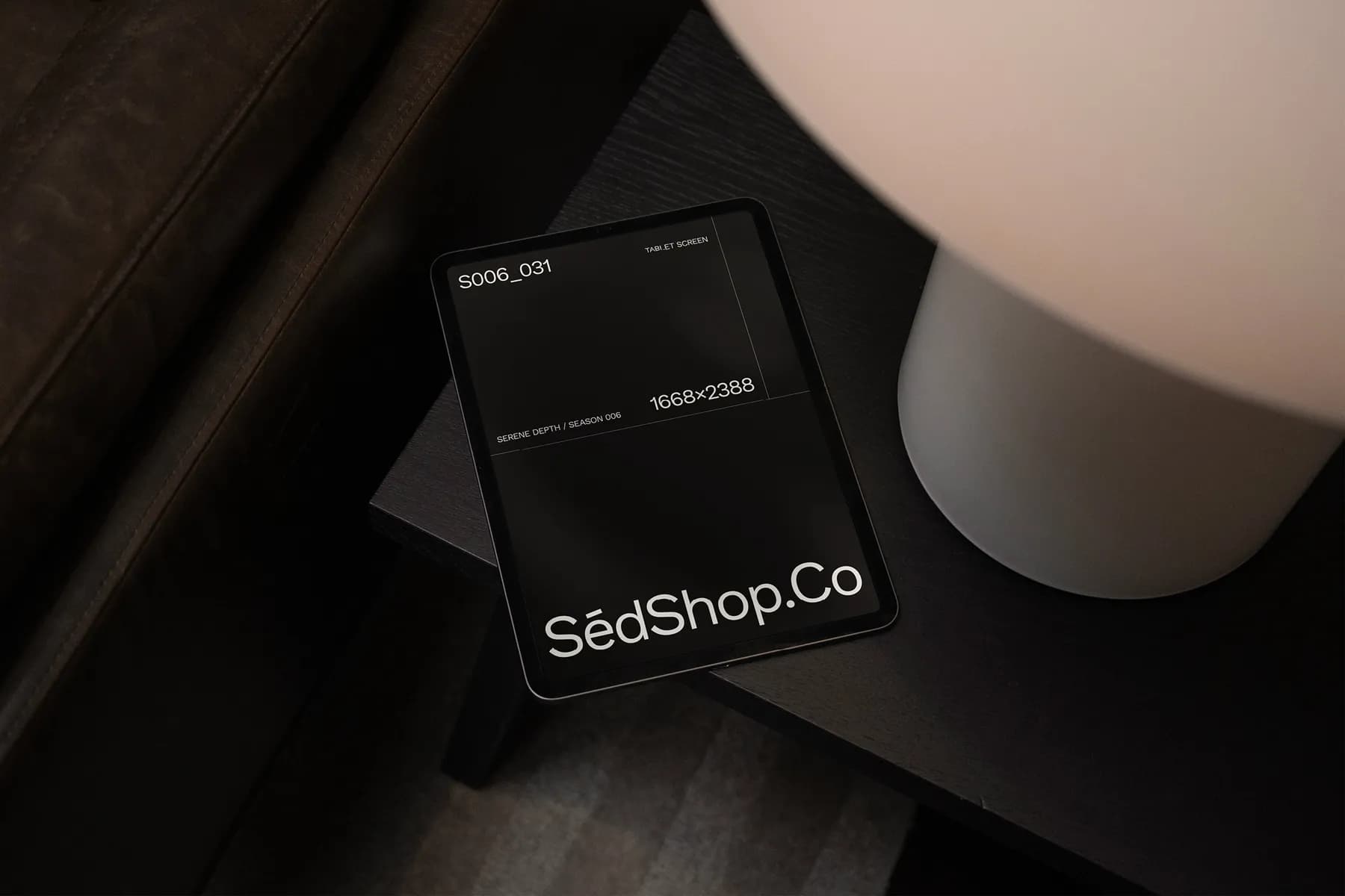 Tablet screen mockup displaying 'SédShop.Co' on a dark interface, placed on a dark table next to a lamp, viewed from an angled perspective.