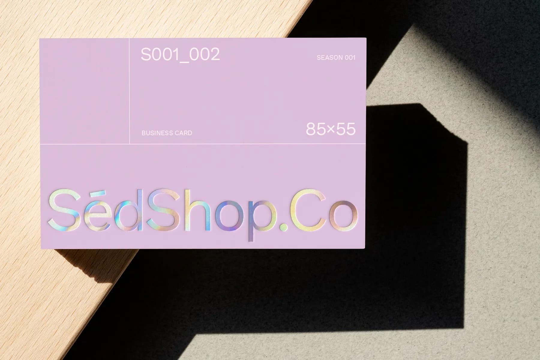Business card mockup featuring a holographic foil effect on a lavender background, placed on a wooden surface with shadows.